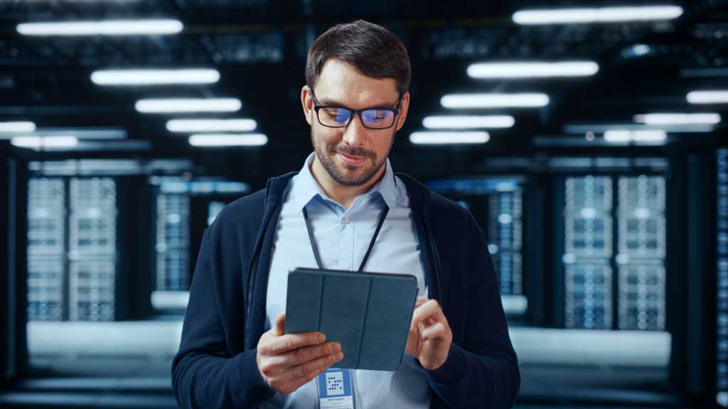 Portrait of a Bearded Handsome Caucasian IT Specialist in Glasses Standing with Tablet and Posing in the Middle of a Working Data Center Server Room with Server Computers Working on a Rack.